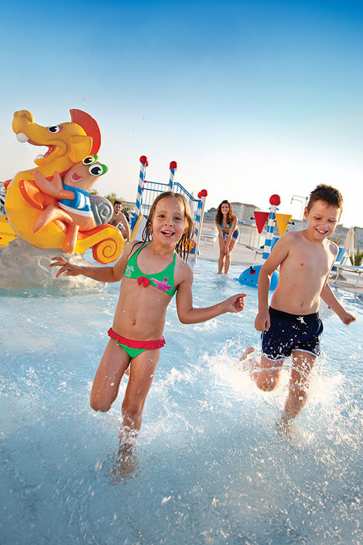 Kinder laufen und spielen im Wasserpark für Kinder, der mit Sprinklern und Wasserrutschen gefüllt ist.