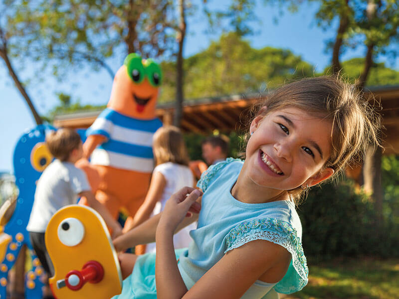 Mädchen lacht mit anderen Kindern und dem Maro-Maskottchen im Hintergrund.