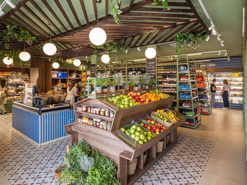 Interno del Valfresco Market con esposizione di frutta fresca, erbe aromatiche, vino e scaffali di generi alimentari, e ospiti che fanno acquisti al campeggio.