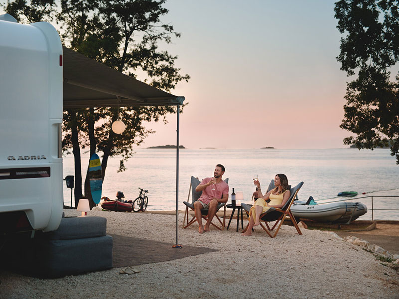 Coppia che si rilassa con un bicchiere di vino in una piazzola Mare sul mare, camper parcheggiato nelle vicinanze, vista sul tramonto.