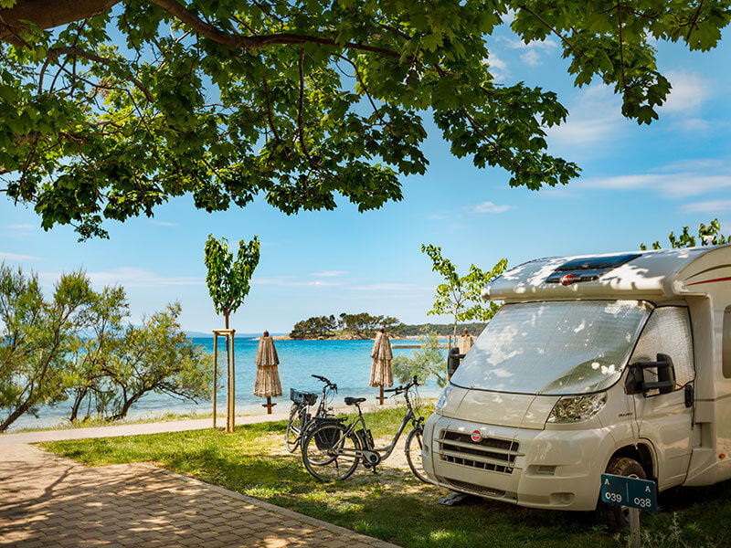 Camper parcheggiato sotto gli alberi alle Padova Camping Pitches con biciclette, mare e ombrelloni sullo sfondo, sull’isola di Rab.