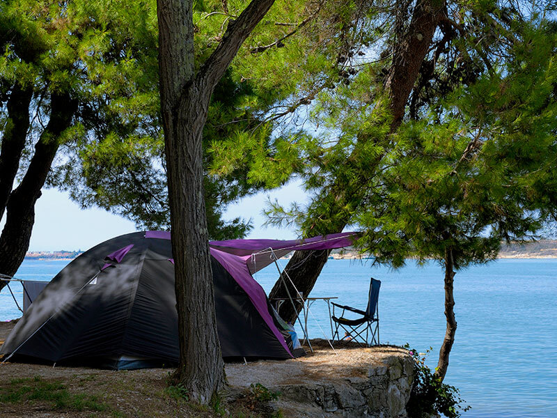 Piazzola per tenda alle Lanterna Camping Pitches, sotto i pini vicino all’acqua, con sedia da campeggio e telo ombreggiante con vista mare.