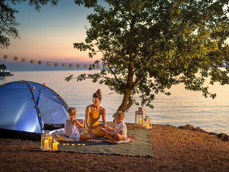 Piazzola per tenda sul mare con una madre e due bambini seduti su una coperta al tramonto, circondati da lanterne e lucine per una serata di campeggio accogliente.