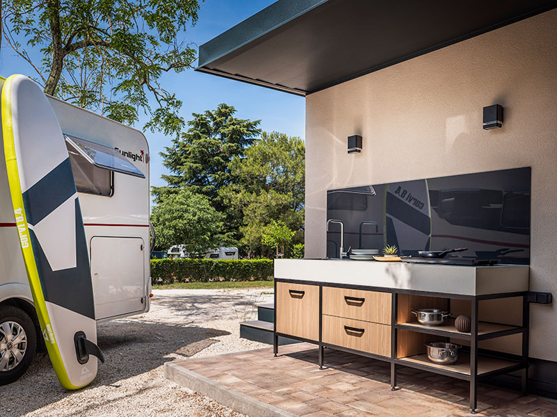 Cucina privata alle Lanterna Camping Pitches con piano di lavoro esterno moderno, cassetti e fornello accanto a un camper e a una tavola da paddle.