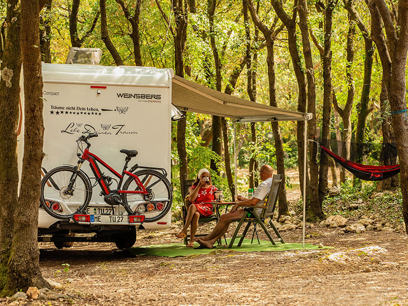 Camper parcheggiato alle Tunarica Camping Pitches in un’area boscosa ombreggiata, con una coppia che si rilassa sotto la veranda e un’amaca sullo sfondo.