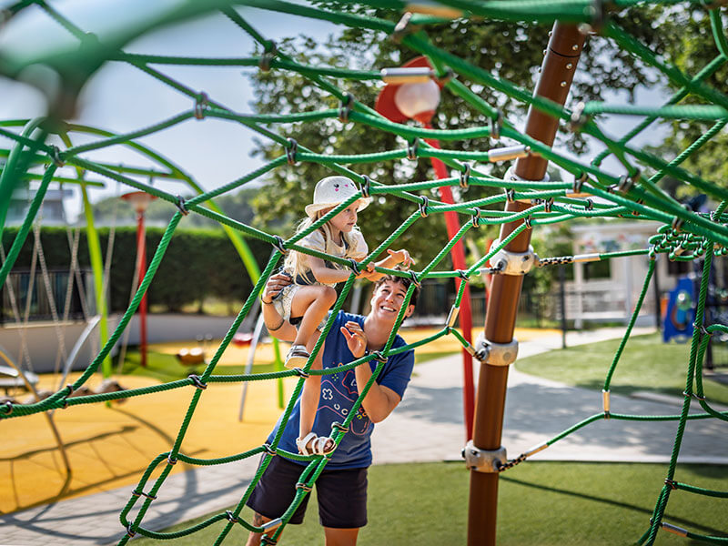Bambino piccolo che si arrampica su una rete di corde in un colorato parco giochi, sotto la supervisione di un adulto, in un campeggio per famiglie.