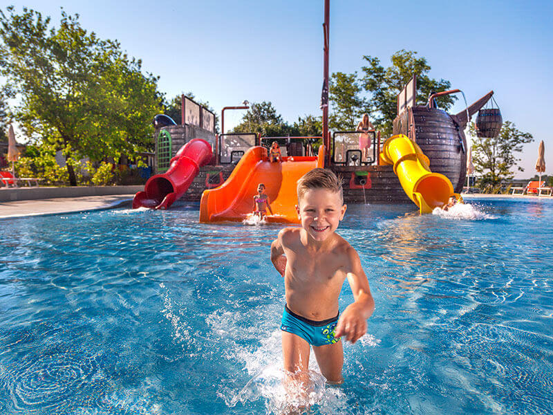 Ragazzo che si diverte in un parco acquatico con scivoli colorati e design a nave pirata nell’area piscine di un campeggio per famiglie.