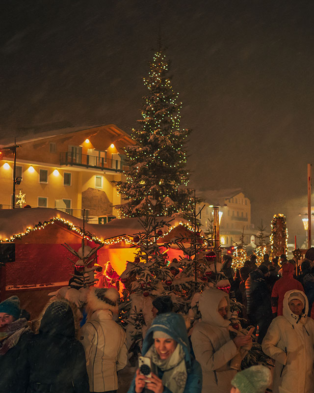 Besucher genießen den festlichen Tauernadvent-Weihnachtsmarkt in Obertauern mit leuchtenden Lichtern, geschmückten Ständen und großem Weihnachtsbaum im Schnee.