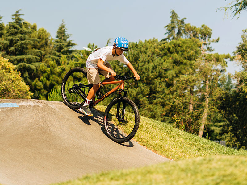 Child in a blue helmet riding an orange bike down a pump track during a bike academy session