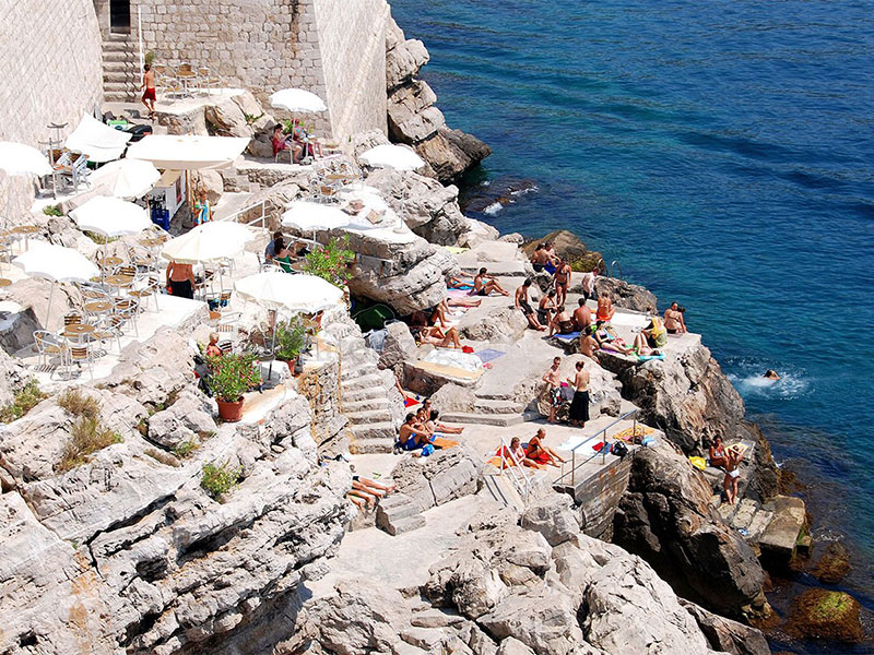 Buža Bar na útesu a plážová zóna s bílými slunečníky a přístupem k moři v Dubrovníku