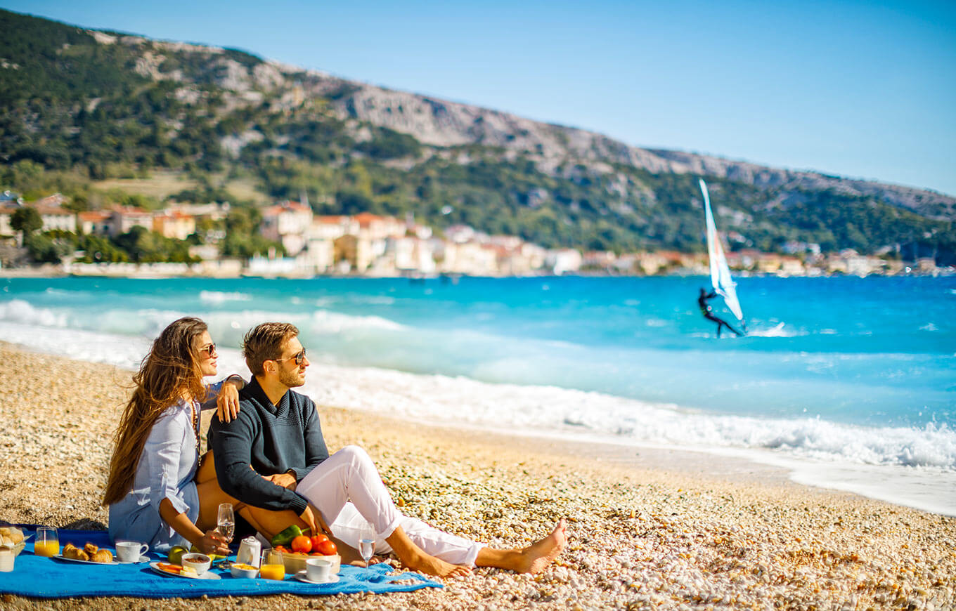 Par se opušta na plaži Vela Plaža s pogledom na jedrilice u Baškoj