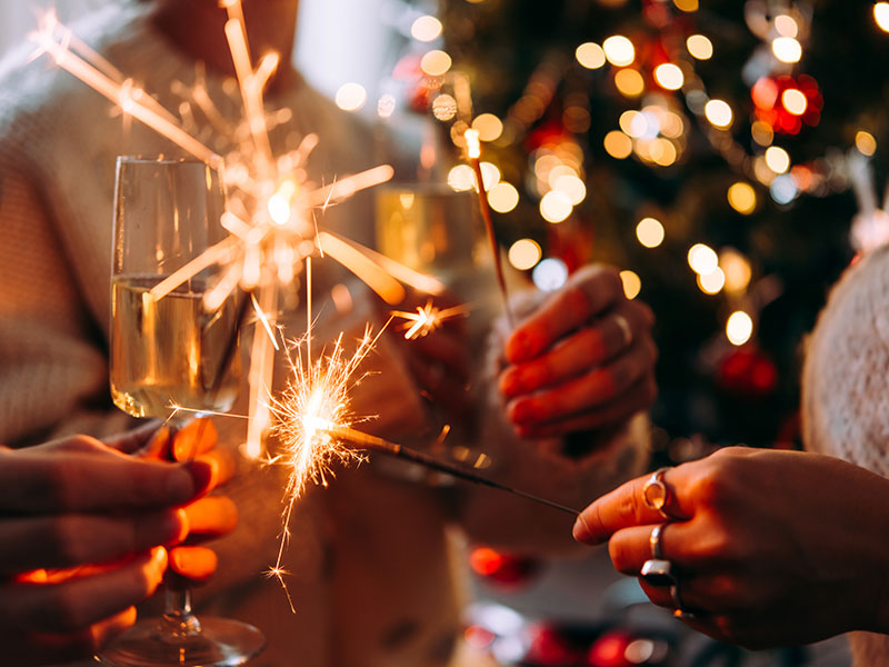 Nahaufnahme von Menschen, die mit Champagnergläsern anstoßen und Wunderkerzen zum Jahreswechsel anzünden, vor dem Weihnachtsbaum.