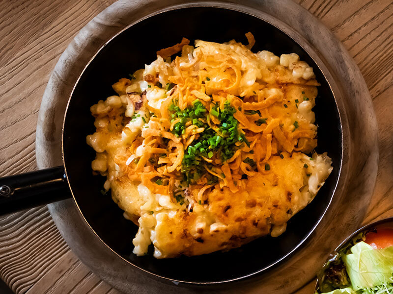 Traditionelle österreichische Käsespätzle, serviert in einer heißen Pfanne, garniert mit knusprigen Röstzwiebeln und frischem Schnittlauch.
