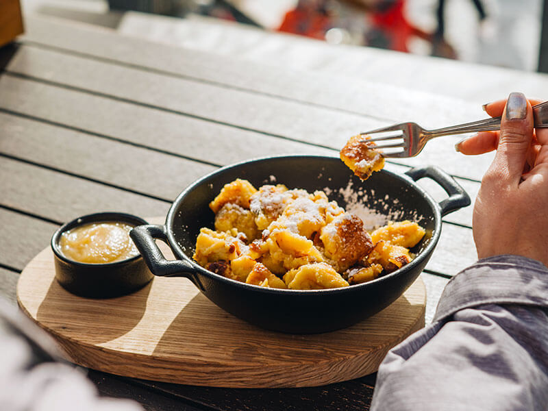 Kaiserschmarrn, ein traditionelles, zerrissenes Pfannkuchendessert, serviert in einer Pfanne mit Apfelmus auf einer sonnigen Alpenterrasse