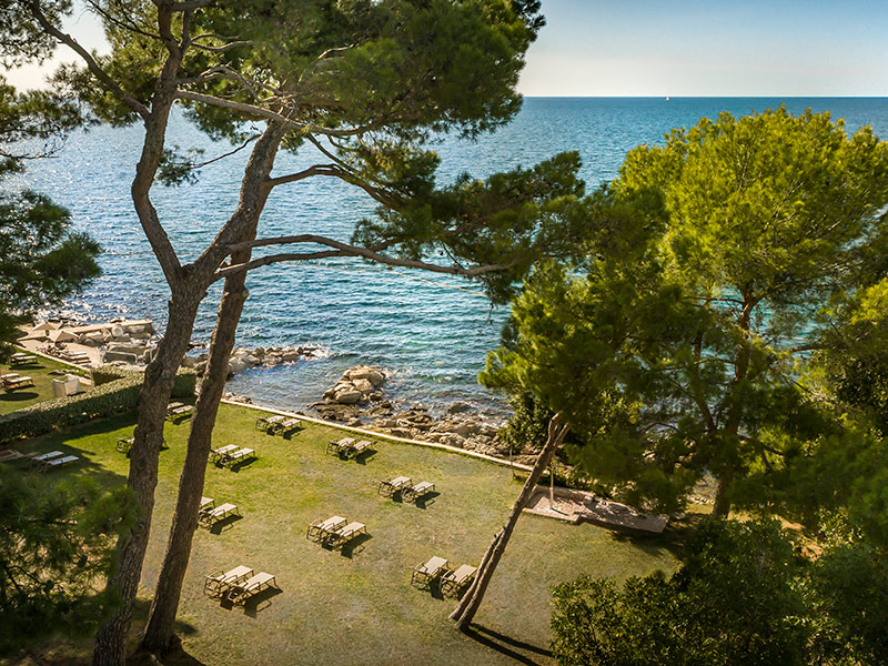 Aerial view of the pebbly Barbaran Beach that has grass spots in the shade with sunbeds and umbrellas