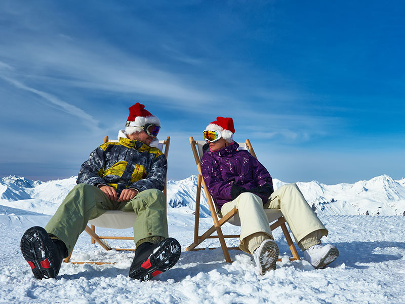 Zwei Skifahrer mit Weihnachtsmützen entspannen auf Liegestühlen im Schnee mit herrlichem Blick auf die Berge in Obertauern.