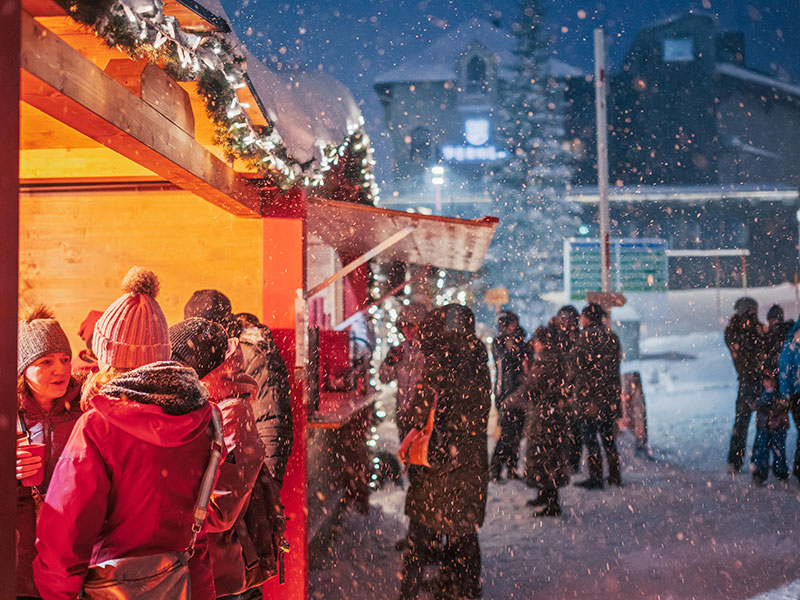 Menschen genießen einen verschneiten Abend auf dem Weihnachtsmarkt in Obertauern, plaudern und trinken an einem warm beleuchteten Holzstand.