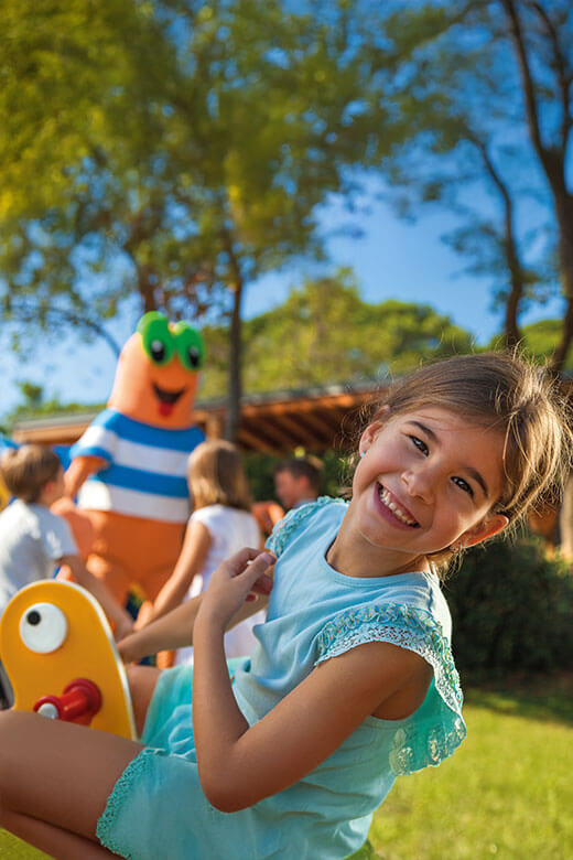 Kleines Mädchen lächelt, während Maro der Krabbe im Hintergrund mit Kindern spielt.