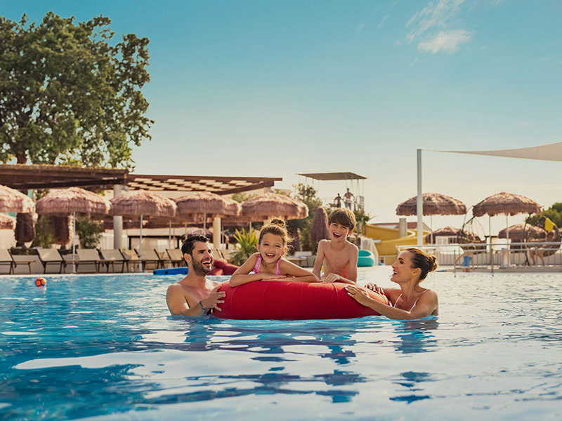 Family of four on a float in an outdoor pool surrounded by lounge chairs and sun umbrellas.