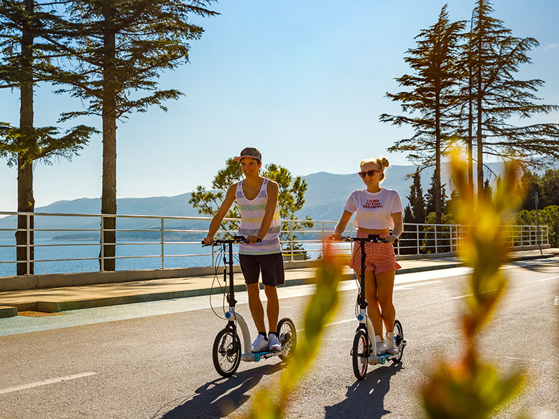 Two teens on scooters riding down a scenic road