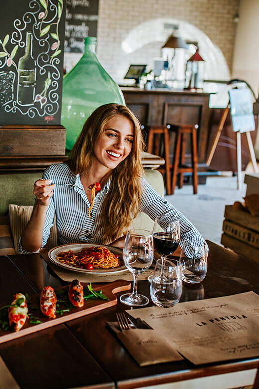 Une femme souriante savourant des pâtes et du vin dans un restaurant chaleureux au décor rustique.