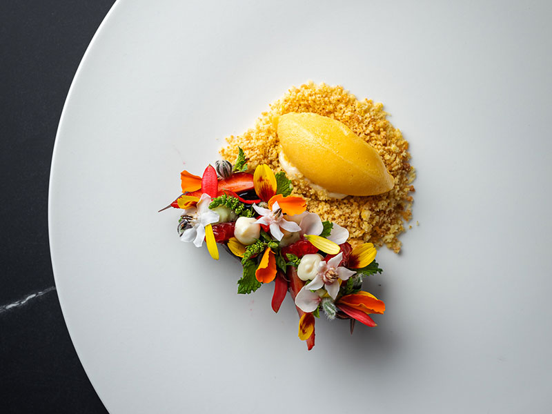 Dessert gastronomique avec sorbet à la mangue et garniture de fleurs comestibles colorées sur une assiette minimaliste.