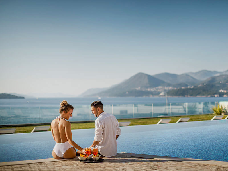 A couple enjoying cocktails and snacks at the edge of a pool with Dubrovnik in the background