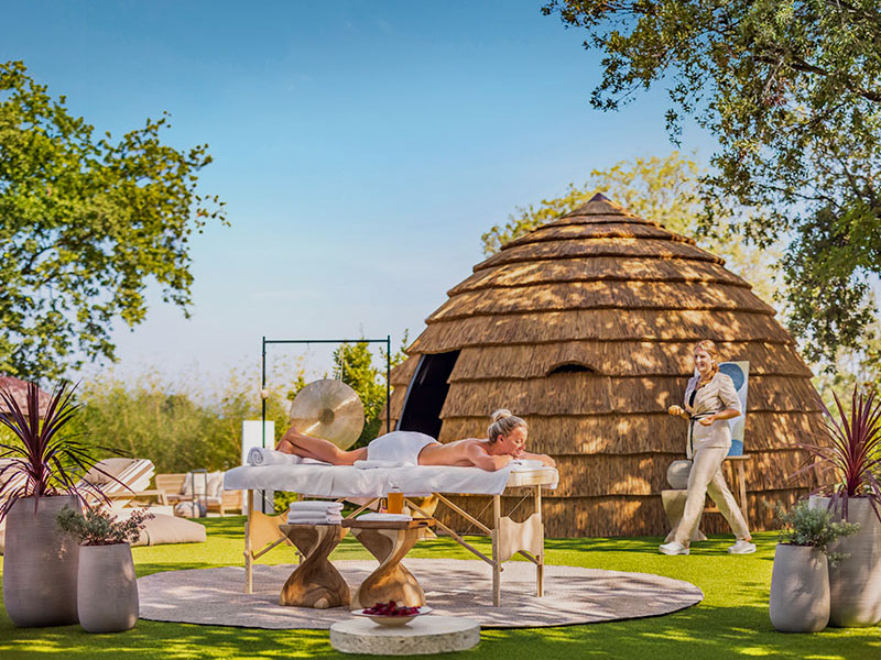 Allestimento per un massaggio rilassante all’aperto vicino a una capanna dal tetto di paglia, circondata dalla natura, in un ambiente spa.