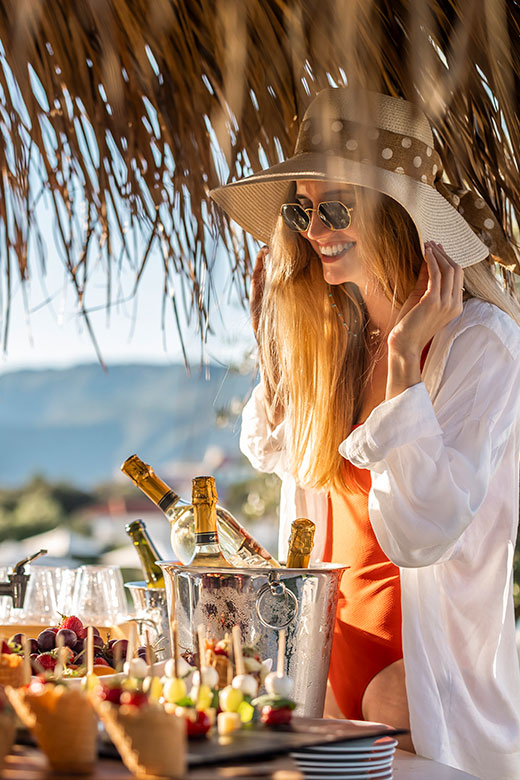 Woman in summer attire enjoying post-spa refreshments with champagne and fruit skewers under a thatched roof