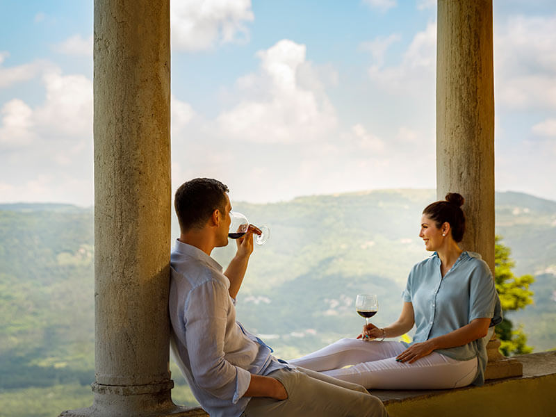 Coppia appoggiata a pilastri, che beve vino e ammira il panorama.